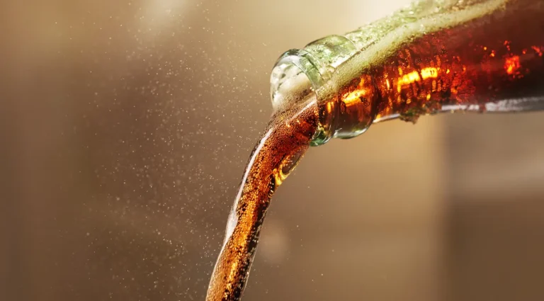 A stream of dark cola pouring into a glass with a soft, blurred beige background.