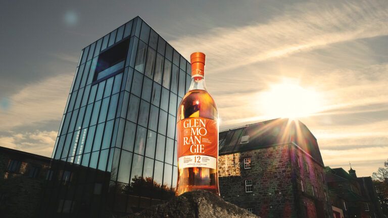 A bottle of Glenmorangie whiskey standing in front of a modern glass office building at sunset.