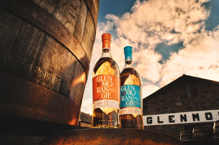 Two bottles of Glenmorangie whiskey standing on a wooden dock by the ocean under a cloudy sky.