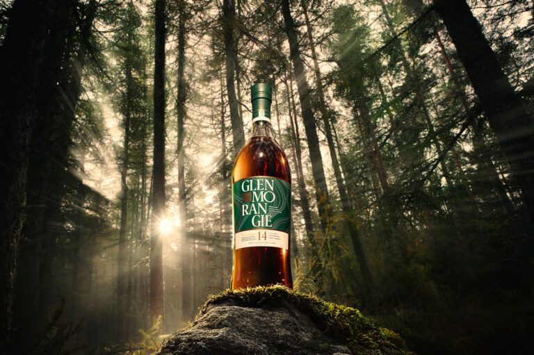 A Glenmorangie whiskey bottle perched on a mossy rock in a misty, sunlit forest.