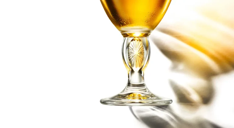 A stemmed Stella Artois chalice filled with golden beer, backlit on a white surface
