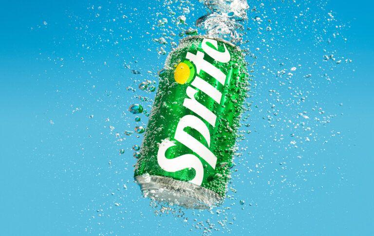 A silver Sprite can exploding through a massive splash of clear water against a blue background.