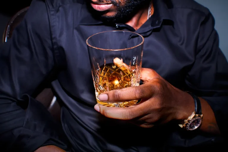 A person in a black shirt holding a glass of whiskey on the rocks in a dark room.