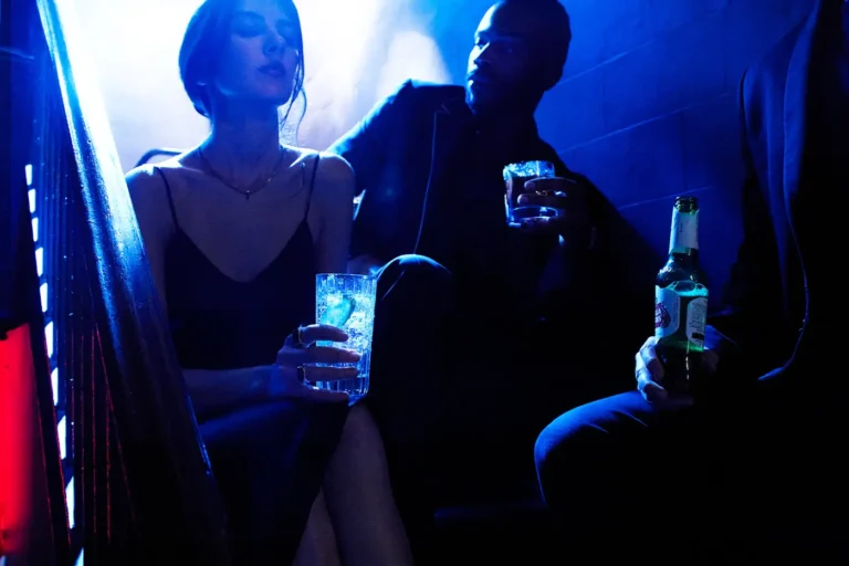 Two people sitting in a dark blue-lit booth at a bar, holding drinks.