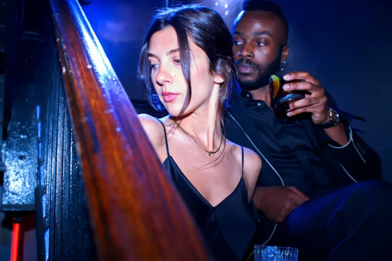 A woman in a black dress holding a drink in a dimly lit, blue-toned nightclub setting.